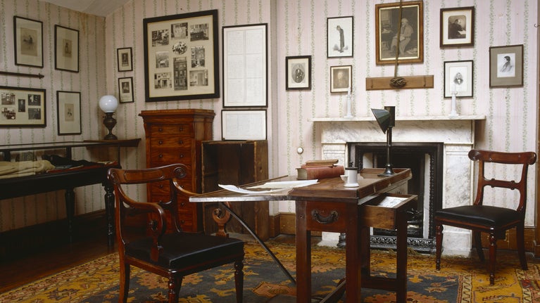 An image of the attic at Carlyle's House in London set out as it was when Carlyle wrote there using it as his study. It is a light room with a writing table and glass display case.
